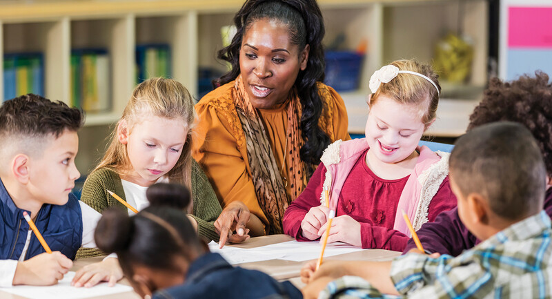 A teacher sitting at a table, pointing to papers, surrounded by students listening and writing 